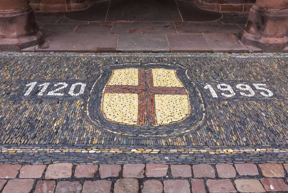 Photo du sol de pribourg où est dessiné un écusson blanc avec une croix rouge. Il y a les nombres "1120" et "1995" de chaque côté de l’écusson.